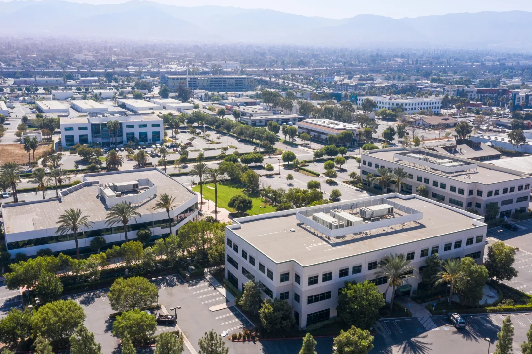Aerial view of a modern business district in Corona, California, featuring office buildings, parking lots, landscaped green spaces, and palm trees under a hazy sky with distant mountains.