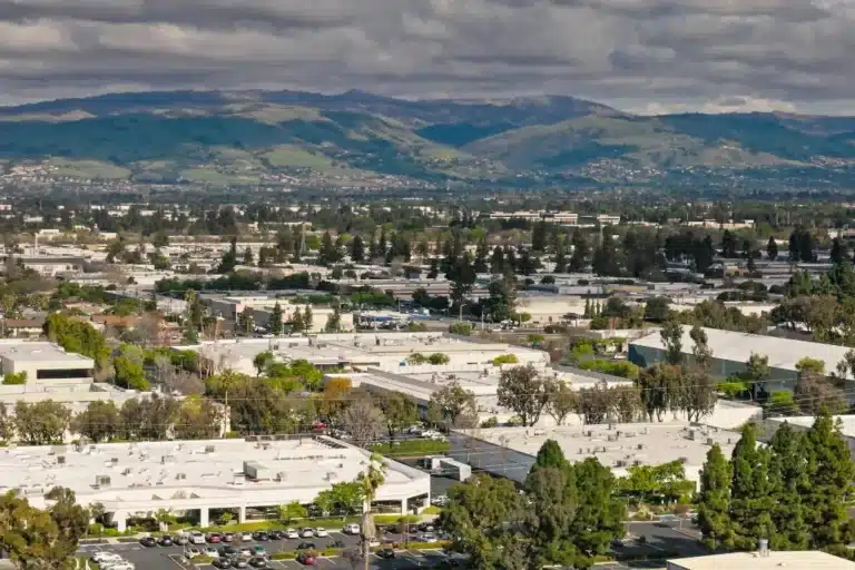 Panoramic cityscape of Santa Clara, California, with low-rise commercial buildings, tree-lined streets, and mountain views under dramatic clouds.
