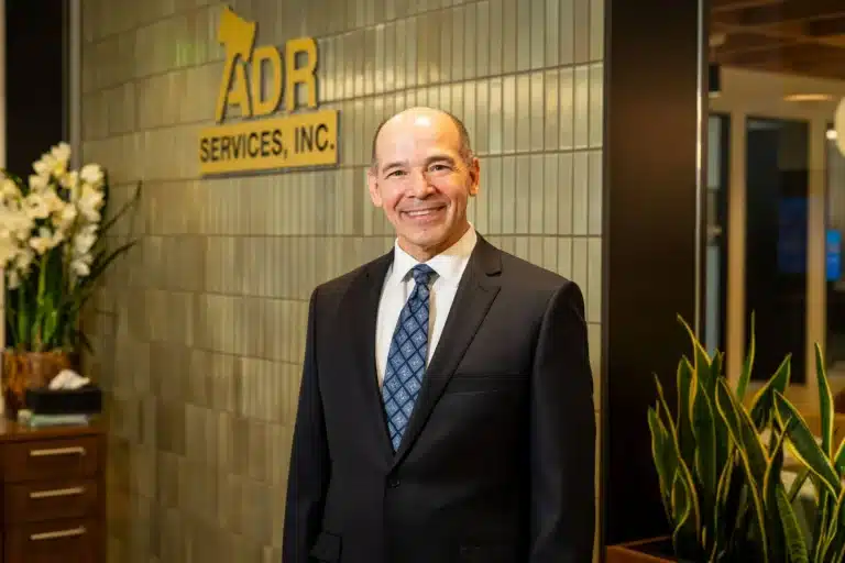 A man in a suit and tie stands confidently in front of a large sign, smiling at the camera.