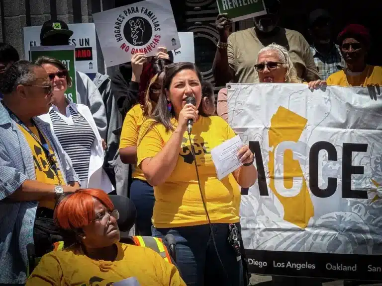 ommunity members rally as a woman speaks into a microphone while protesters hold housing justice signs.